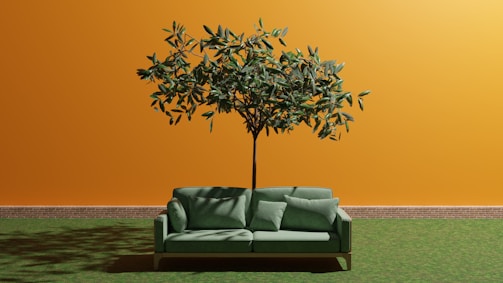 A green sofa with cushions is positioned in front of an orange wall with a tree growing through the center of the sofa. The wall has a brick baseboard, and the ground is covered with grass, creating an unusual and artistic indoor-outdoor blend.