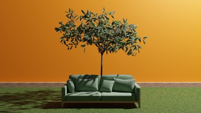A green sofa with cushions is positioned in front of an orange wall with a tree growing through the center of the sofa. The wall has a brick baseboard, and the ground is covered with grass, creating an unusual and artistic indoor-outdoor blend.