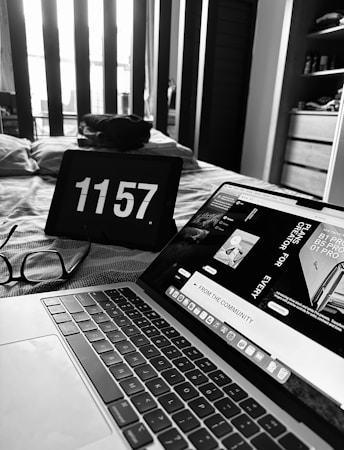 A grayscale image of a cozy bedroom with a modern aesthetic. A laptop is open displaying a webpage, and an iPad nearby shows the time 11:57. Glasses rest on the bedspread, which has a subtle pattern. Light streams in through the window, casting soft shadows across the room.