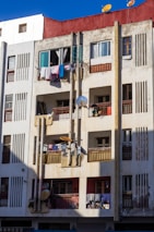 A multi-story residential building with a red top section, featuring numerous balconies. Laundry is hanging to dry on several balconies. There are multiple satellite dishes attached to the building's facade, and the structure appears weathered with some visible dirt and wear.