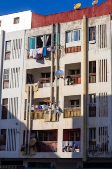 A multi-story residential building with a red top section, featuring numerous balconies. Laundry is hanging to dry on several balconies. There are multiple satellite dishes attached to the building's facade, and the structure appears weathered with some visible dirt and wear.