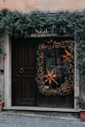 A rustic wooden door is adorned with a large holiday wreath decorated with warm, glowing lights and a prominent star-shaped ornament. Ivy climbs and cascades around the doorframe and sign, adding a natural touch. The sign above reads 'Bed & Breakfast' blending into the cozy and welcoming atmosphere. Stone tiles lead up to the entrance, enhancing the quaint charm.
