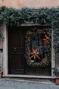 A rustic wooden door is adorned with a large holiday wreath decorated with warm, glowing lights and a prominent star-shaped ornament. Ivy climbs and cascades around the doorframe and sign, adding a natural touch. The sign above reads 'Bed & Breakfast' blending into the cozy and welcoming atmosphere. Stone tiles lead up to the entrance, enhancing the quaint charm.