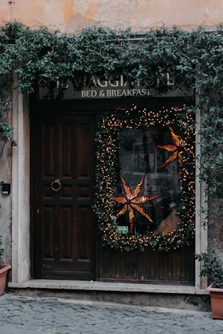 A rustic wooden door is adorned with a large holiday wreath decorated with warm, glowing lights and a prominent star-shaped ornament. Ivy climbs and cascades around the doorframe and sign, adding a natural touch. The sign above reads 'Bed & Breakfast' blending into the cozy and welcoming atmosphere. Stone tiles lead up to the entrance, enhancing the quaint charm.