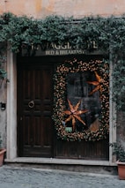 A rustic wooden door is adorned with a large holiday wreath decorated with warm, glowing lights and a prominent star-shaped ornament. Ivy climbs and cascades around the doorframe and sign, adding a natural touch. The sign above reads 'Bed & Breakfast' blending into the cozy and welcoming atmosphere. Stone tiles lead up to the entrance, enhancing the quaint charm.