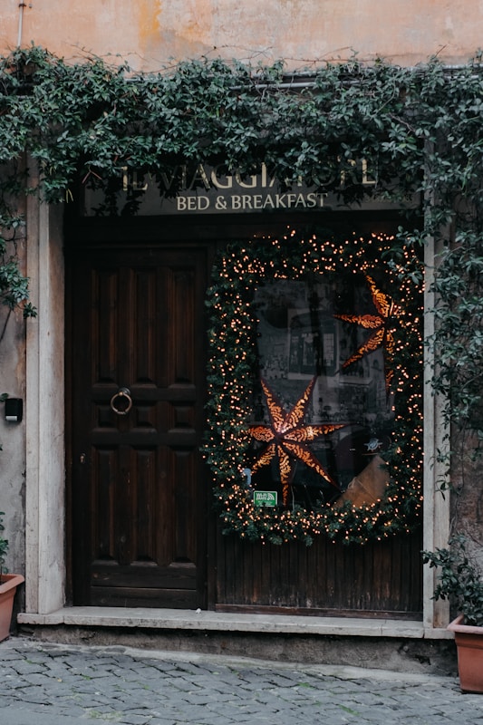 A rustic wooden door is adorned with a large holiday wreath decorated with warm, glowing lights and a prominent star-shaped ornament. Ivy climbs and cascades around the doorframe and sign, adding a natural touch. The sign above reads 'Bed & Breakfast' blending into the cozy and welcoming atmosphere. Stone tiles lead up to the entrance, enhancing the quaint charm.