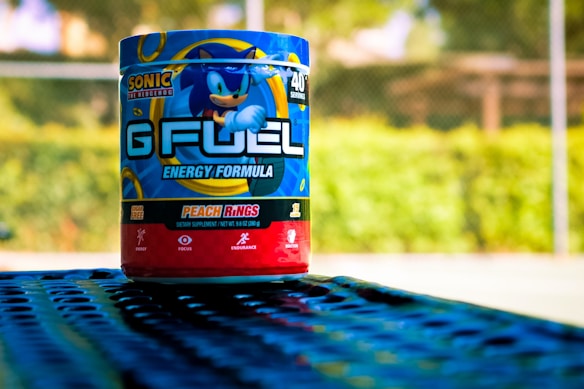 A container of G FUEL energy powder featuring Sonic the Hedgehog on the label is placed on a perforated blue metal surface. The product is identified as the 'Energy Formula' flavor 'Peach Rings,' and it is labeled as sugar-free. The background consists of blurred green foliage and what appears to be a tennis court fence.