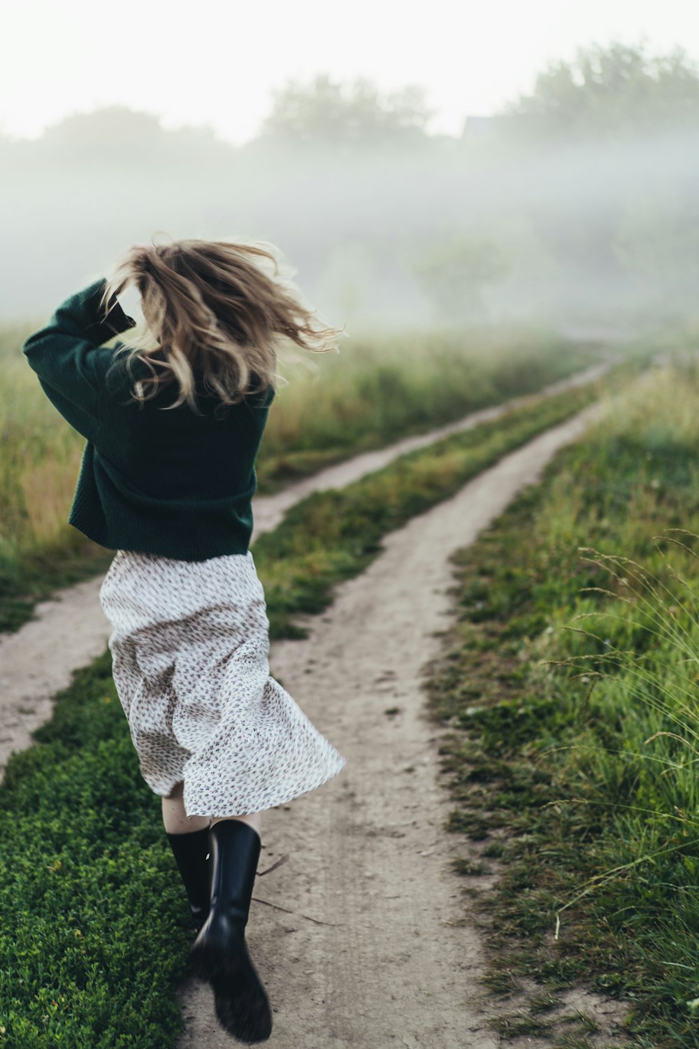 a woman walking down a dirt road next to a lush green field