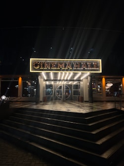 An illuminated sign above a cinema entrance, displaying the words 'CINEMA HALL' in bright lights. The entrance features a set of closed glass doors, flanked by lit glass windows, with a series of wide steps leading up to it. The warm glow from the sign and interior lights contrasts against the surrounding dark night.