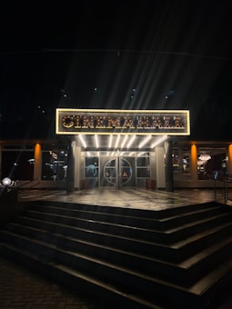 An illuminated sign above a cinema entrance, displaying the words 'CINEMA HALL' in bright lights. The entrance features a set of closed glass doors, flanked by lit glass windows, with a series of wide steps leading up to it. The warm glow from the sign and interior lights contrasts against the surrounding dark night.