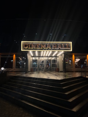 An illuminated sign above a cinema entrance, displaying the words 'CINEMA HALL' in bright lights. The entrance features a set of closed glass doors, flanked by lit glass windows, with a series of wide steps leading up to it. The warm glow from the sign and interior lights contrasts against the surrounding dark night.