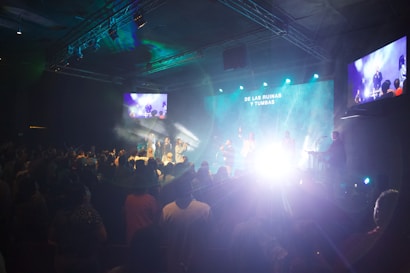 A live performance is taking place in a dimly lit indoor space. The room is filled with people watching the stage, where several performers are illuminated by bright lights against a blue and green backdrop. There are screens on the sides showing the stage activity. Smoke or fog effects enhance the dramatic lighting on stage.