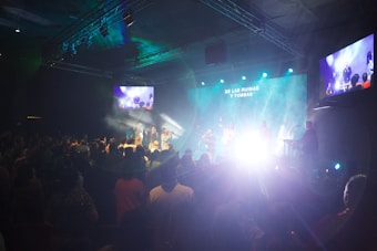 A live performance is taking place in a dimly lit indoor space. The room is filled with people watching the stage, where several performers are illuminated by bright lights against a blue and green backdrop. There are screens on the sides showing the stage activity. Smoke or fog effects enhance the dramatic lighting on stage.