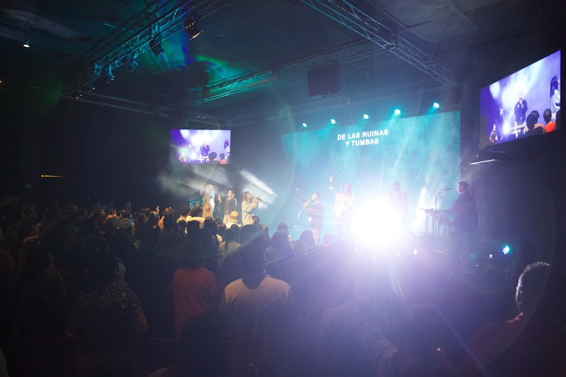 A live performance is taking place in a dimly lit indoor space. The room is filled with people watching the stage, where several performers are illuminated by bright lights against a blue and green backdrop. There are screens on the sides showing the stage activity. Smoke or fog effects enhance the dramatic lighting on stage.