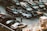 A parking lot features multiple levels of cars, with vehicles parked on elevated platforms. A person stands near a white car in the foreground, adding a sense of scale and activity to the scene. The area is busy with various types of cars, indicating a possible urban setting.