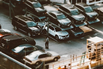 A parking lot features multiple levels of cars, with vehicles parked on elevated platforms. A person stands near a white car in the foreground, adding a sense of scale and activity to the scene. The area is busy with various types of cars, indicating a possible urban setting.