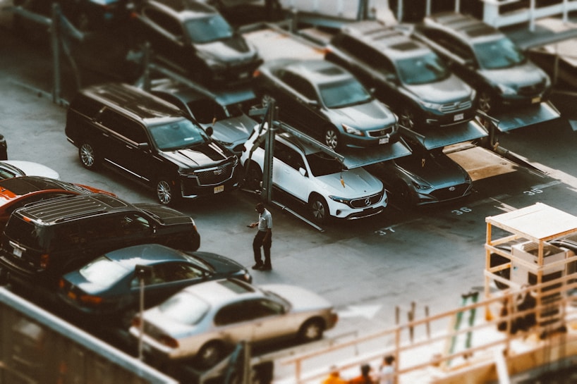 A parking lot features multiple levels of cars, with vehicles parked on elevated platforms. A person stands near a white car in the foreground, adding a sense of scale and activity to the scene. The area is busy with various types of cars, indicating a possible urban setting.