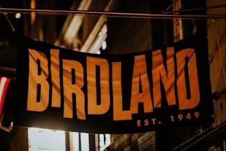 A sign with large bold letters spells out 'BIRDLAND' along with the text 'EST. 1949' beneath it. The sign appears to be hanging in an urban setting with dim lighting, possibly outside of a building.