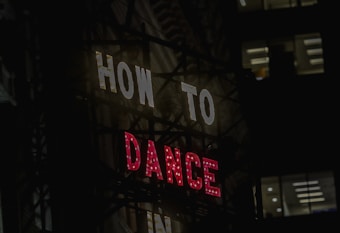 A neon sign with the words 'HOW TO' in white and 'DANCE' in red, set against a dark urban backdrop. Windows of a nearby building are visible in the background.
