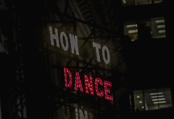 A neon sign with the words 'HOW TO' in white and 'DANCE' in red, set against a dark urban backdrop. Windows of a nearby building are visible in the background.