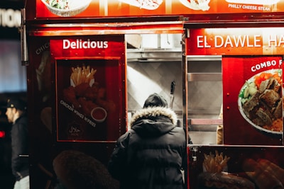 A person in a black, fur-lined hooded coat stands at a food truck that features bright red signage. The signs display images of various dishes such as fried chicken wings, fries, and a combo platter. Words like 'Delicious' and 'Halal' are prominent, indicating the type of food served.