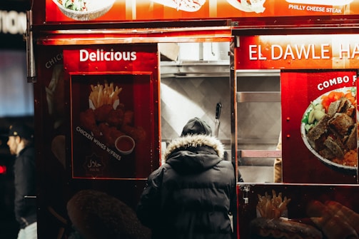A person in a black, fur-lined hooded coat stands at a food truck that features bright red signage. The signs display images of various dishes such as fried chicken wings, fries, and a combo platter. Words like 'Delicious' and 'Halal' are prominent, indicating the type of food served.