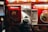 A person in a black, fur-lined hooded coat stands at a food truck that features bright red signage. The signs display images of various dishes such as fried chicken wings, fries, and a combo platter. Words like 'Delicious' and 'Halal' are prominent, indicating the type of food served.