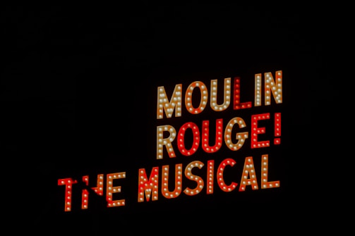 Brightly lit sign with the words 'Moulin Rouge! The Musical' against a black background. The lettering is bold and illuminated with lights.