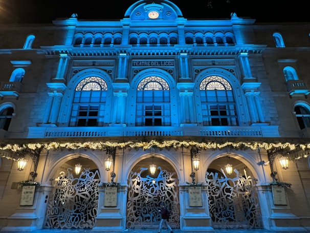 A grand architectural facade featuring arched windows and ornate decorations, illuminated with blue lighting. The entrance displays intricate wrought iron designs and festive garlands are adorned on the structure. Several classical-style lamps enhance the elegant aesthetic.