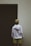 A person wearing a white hoodie with bold black text stands facing a dark, closed garage door. The person also wears a beige cap and lime green shorts. The text on the hoodie reads 'Breezy Even When The Coast Is Uneasy.' The surrounding wall is light-colored, creating a stark contrast with the dark door.