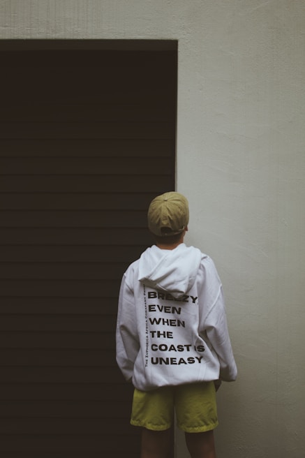 A person wearing a white hoodie with bold black text stands facing a dark, closed garage door. The person also wears a beige cap and lime green shorts. The text on the hoodie reads 'Breezy Even When The Coast Is Uneasy.' The surrounding wall is light-colored, creating a stark contrast with the dark door.