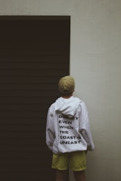 A person wearing a white hoodie with bold black text stands facing a dark, closed garage door. The person also wears a beige cap and lime green shorts. The text on the hoodie reads 'Breezy Even When The Coast Is Uneasy.' The surrounding wall is light-colored, creating a stark contrast with the dark door.