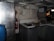 A dimly lit industrial kitchen with a metal refrigerator, a sink with two basins, and a fire extinguisher on the wall. The walls are painted white, and there is a large vent or exhaust hood above the sink area. The setting appears worn and utilitarian.