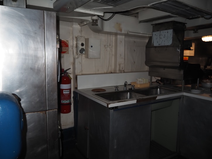 A dimly lit industrial kitchen with a metal refrigerator, a sink with two basins, and a fire extinguisher on the wall. The walls are painted white, and there is a large vent or exhaust hood above the sink area. The setting appears worn and utilitarian.