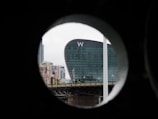 A modern, curved glass building with a large 'W' logo is seen through a circular frame. Surrounding the main building are various other buildings and a bridge in an urban setting.