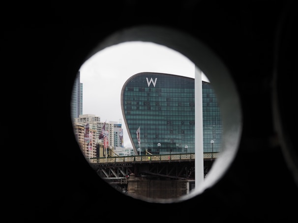 A modern, curved glass building with a large 'W' logo is seen through a circular frame. Surrounding the main building are various other buildings and a bridge in an urban setting.