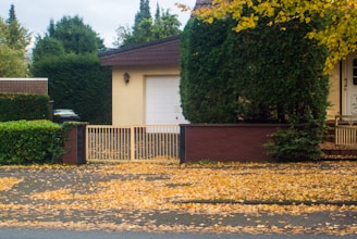 A suburban setting featuring a house with a closed white garage door and a fence with a gate. Trees and bushes with lush green foliage surround the area, and the ground is covered in fallen yellow leaves, indicating an autumn season.