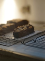 A chocolate brownie topped with almond slices rests on a wire cooling rack. The image has a soft, warm light coming from the background, creating a cozy atmosphere.
