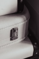 A close-up view of a textured white suitcase with metal hardware, featuring a latch and accentuated by black straps on the sides.