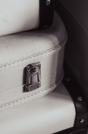 A close-up view of a textured white suitcase with metal hardware, featuring a latch and accentuated by black straps on the sides.