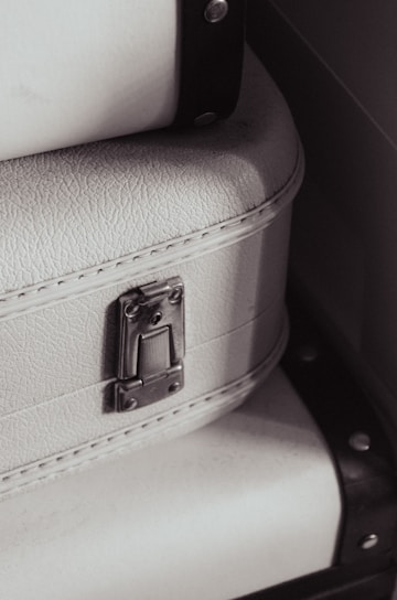 A close-up view of a textured white suitcase with metal hardware, featuring a latch and accentuated by black straps on the sides.