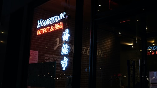 A dimly lit restaurant exterior with neon signs displaying the words 'Hometown Hotpot & BBQ'. The signage includes both English and Chinese characters, illuminated in blue and red against a backdrop of a brick wall.