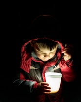 A young child wearing a winter jacket with a hood is holding a white cup while shining a flashlight into it. The child&rsquo;s face is softly illuminated by the light, creating a cozy atmosphere against a dark background.