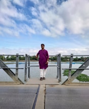 A person wearing a purple kurta and white pants stands on a metal walkway over a body of water. The sky is filled with clouds, and green vegetation is visible in the background. The individual is wearing sunglasses and black sandals.