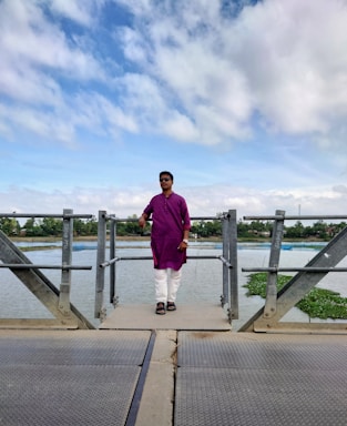 A person wearing a purple kurta and white pants stands on a metal walkway over a body of water. The sky is filled with clouds, and green vegetation is visible in the background. The individual is wearing sunglasses and black sandals.