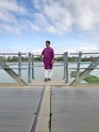 A person wearing a purple kurta and white pants is standing on a metal bridge over a body of water. The sky is partly cloudy, and there is greenery in the distance.