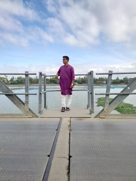 A person wearing a purple kurta and white pants is standing on a metal bridge over a body of water. The sky is partly cloudy, and there is greenery in the distance.