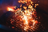 A barbecue grill with glowing charcoal briquettes emits bright, fiery sparks. The grill bars are illuminated by the intense heat of the coals, creating a dynamic scene with orange and yellow flames contrasting against the dark background.
