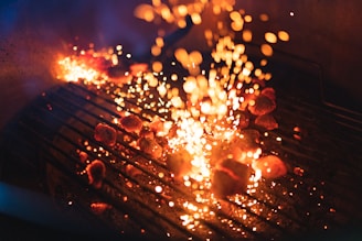 A barbecue grill with glowing charcoal briquettes emits bright, fiery sparks. The grill bars are illuminated by the intense heat of the coals, creating a dynamic scene with orange and yellow flames contrasting against the dark background.