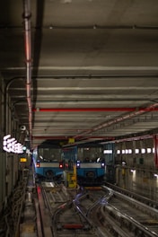 Two blue metro trains are parked side by side on separate tracks inside a dimly lit underground subway tunnel. The tunnel features a concrete ceiling with red and black piping, illuminated directional signs on the left, and glowing lights on the right wall. Tracks and various technical installations are visible on the floor.
